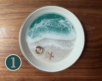 May include: A round white ceramic trinket dish featuring a resin art design of a beach scene. The design includes a teal and white wave pattern, a sandy base with embedded glitter, a small gold-toned seashell with a pearl, and a gold starfish.