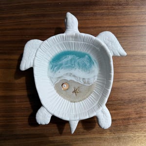 May include: White ceramic turtle-shaped trinket dish with a beach scene inside. The dish features a turquoise and white wave design, sand, a pearl, and a starfish. The dish is on a wooden surface.