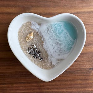 May include: White heart-shaped dish with a blue and white resin ocean scene. The scene includes a seashell and a silver lobster charm.