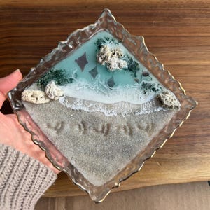 May include: A clear, diamond-shaped resin tray with a gold-colored trim. The tray features a beach scene with a sandy shore, turquoise water, and coral accents. The tray has a gold-colored trim.