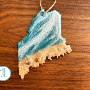 May include: A blue and white resin ornament shaped like the state of Maine. The ornament has a sandy bottom with gold glitter accents.