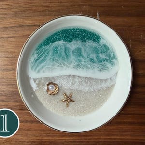 Op de afbeelding: Een ronde witte keramische schaal met een hars-oceaanlandschap met blauwgroen glitterwater, witte schuimgolven en een zandstrand. De schaal is versierd met een kleine goudkleurige schelp met een witte parel en een gouden zeester.
