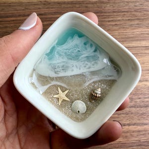 May include: A white square dish with a resin ocean scene. The scene includes blue water with white foam, tan sand, a starfish, a seashell, and a small white seashell.