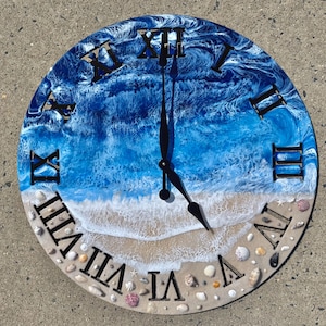 Peut inclure: Horloge murale ronde avec un motif de plage. Le cadran de l'horloge présente un motif de vagues océaniques bleues et blanches, avec des chiffres romains noirs et des aiguilles. Le bord extérieur est décoré de coquillages.