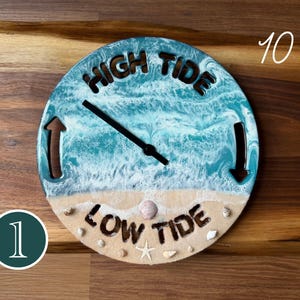 Pode incluir: Um relógio de marés redondo de 25,4 cm com uma cena oceânica em resina, ondas brancas, uma praia de areia e pequenas conchas. O mostrador exibe o texto HIGH TIDE no topo e LOW TIDE na base, com um ponteiro preto e setas direcionais.