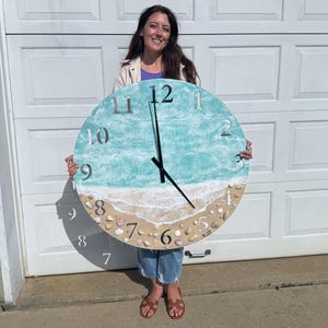 May include: Large, round wall clock with a beach-themed design. The clock face features a turquoise and white ocean scene with a sandy beach at the bottom. Silver numerals mark the hours, and the clock hands are black.