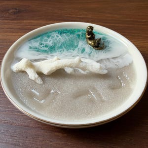 Puede incluir: Un plato decorativo redondo de cerámica con una escena de playa hecha en resina. El diseño incluye una base color arena, una rama de coral blanca, un patrón de olas en color verde azulado y blanco, y una pequeña figura de nutria bronce.