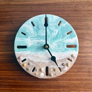 May include: Round wall clock with a beach-themed design. The clock face features a turquoise and white resin ocean scene with embedded seashells and sand. Black clock hands point to the time. The clock is mounted on a wooden surface.