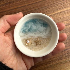 Peut inclure: Un plat en céramique blanche avec une scène océanique en résine bleue et blanche. Le plat contient un petit coquillage doré avec une perle à l'intérieur et une étoile de mer dorée.