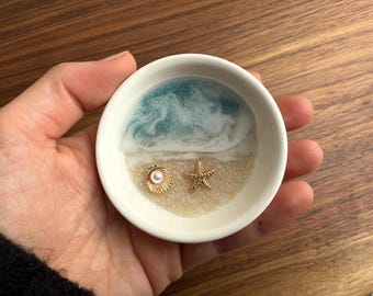 May include: A white ceramic dish with a blue and white resin ocean scene. The dish has a small golden seashell with a pearl inside and a golden starfish.