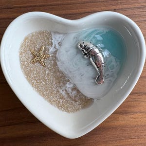 Puede incluir: Plato de cerámica blanco en forma de corazón con un diseño de resina azul y blanco. El diseño de resina presenta un manatí y una estrella de mar. El plato es perfecto para guardar joyas u otros objetos pequeños.