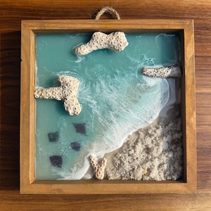 May include: A framed resin art piece depicting an ocean scene. The artwork features a turquoise ocean with white waves, beige sand, and coral accents. The frame is made of natural wood, and the piece is designed to be hung.