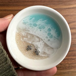 Op de afbeelding: Witte keramische ringenschotel met een blauw en wit harsontwerp dat lijkt op een oceaan golf en strand. Twee zilveren schildpadcharms bevinden zich op het zand.