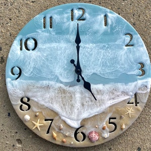 Può includere: Un orologio rotondo con un design a onde oceaniche blu e bianche. Il quadrante dell'orologio ha una spiaggia sabbiosa con conchiglie e una stella marina. I numeri da 1 a 12 sono visualizzati sul quadrante dell'orologio.