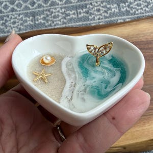 May include: A heart-shaped white ceramic trinket dish with a beach design. It has a sandy area with a gold starfish and shell, and a turquoise resin ocean with a gold whale tail. The dish is held in a hand.
