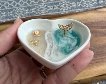 May include: A heart-shaped white ceramic trinket dish with a beach-themed design. It features a sandy area with a gold starfish and shell, and a turquoise resin ocean with a gold whale tail. The dish is held in a hand.