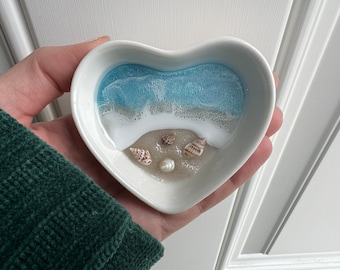 May include: A white heart-shaped dish with a blue and white resin ocean scene and seashells.