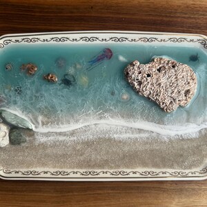 May include: A rectangular decorative tray with a beach scene. The tray features a sandy beach, a turquoise ocean with marine life, and a large, textured rock. The tray has a white border with a gold filigree design.