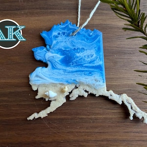 May include: A resin ornament in the shape of Alaska, featuring a blue and white wave design at the top and a sandy beach with a seashell at the bottom. The ornament hangs from a white string.