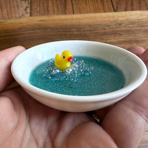 Pode incluir: Um pequeno prato de cerâmica branca cheio de glitter azul-turquesa e contas transparentes. Um patinho de borracha amarelo com bico vermelho flutua no centro. O prato é segurado em uma mão.