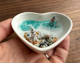 May include: A white heart-shaped dish featuring a miniature seascape scene. The scene includes a lighthouse, a sailboat, and small colourful stones on a blue and white resin base.