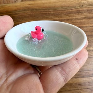 May include: A small, white ceramic dish filled with a shimmering, light blue liquid. A pink flamingo figurine with a black beak floats in the liquid, surrounded by small, clear beads. The dish is held in a hand.
