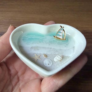 Peut inclure: Un plat en céramique blanc en forme de cœur avec une scène de plage miniature. La scène comprend un océan en résine turquoise, un petit voilier, du sable, une étoile de mer et des coquillages. Le plat est tenu dans une main.