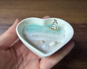 May include: A heart-shaped, white ceramic dish with a miniature beach scene. The scene includes a turquoise resin ocean, a small sailboat, sand, a starfish, and seashells. The dish is held in a hand.