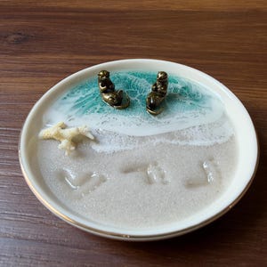 Puede incluir: Un plato decorativo redondo de cerámica blanca con una escena oceánica de resina, dos pequeñas figuras de nutrias de bronce, un trozo de coral blanco y una superficie de resina color arena con huellas y corazones.