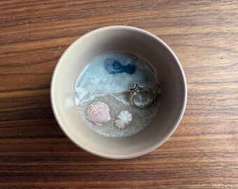 May include: A beige ceramic bowl with a ring, two seashells, and a blue and white resin design that resembles a beach scene.