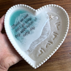 Puede incluir: Plato decorativo en forma de corazón con un diseño de playa. El plato presenta una escena oceánica de resina turquesa y blanca con una estrella de mar y una concha. El texto "Aquellos a quienes amamos no se van, caminan a nuestro lado todos los días" está escrito en el plato.