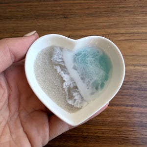 Może przedstawiać: Biały ceramiczny talerz w kształcie serca z motywem plażowym. Talerz ma piaszczysty obszar po lewej stronie i turkusowy wzór oceanu po prawej stronie, z białymi detalami fal. Talerz jest trzymany w dłoni.