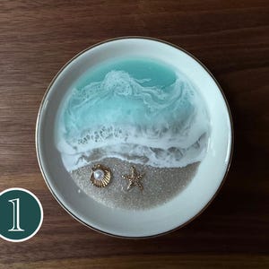 Op de afbeelding: Een ronde witte keramische schaal met een harsontwerp dat een strandscène nabootst. Het ontwerp bevat een zandkleurige basis met een goudkleurige schelp met een witte parel en een goudkleurige zeester, overgoten met turquoise gekleurd water.