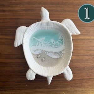 May include: White ceramic turtle-shaped trinket dish with a beach scene inside. The dish features a turquoise and white wave design, sand, and two small silver turtle figurines. The number "1" is in a teal circle in the upper right corner.