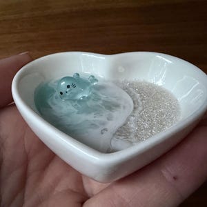 Può includere: Un piattino in ceramica bianco a forma di cuore con un inserto in resina che imita una spiaggia. Il design include una piccola statuetta di foca traslucida azzurra che riposa tra onde in resina blu e bianca accanto a della sabbia bianca.