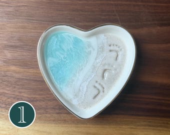 May include: A heart-shaped ceramic trinket dish featuring a resin design that mimics a beach scene. The left side displays swirling light blue and white ocean waves, while the right side shows textured beige sand with two small footprints imprinted.