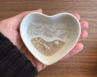 Op de afbeelding: Witte hartvormige schaal met een strandscene erin. De scène bevat witte schuimende golven, bruin zand, een zeester, een schelp en een kleine schelp.