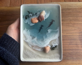 May include: Rectangular resin tray with a coastal scene. The tray features a light blue and white ocean design with three hammerhead sharks, sand, and small rocks. The tray has a gold trim and is held by a person.