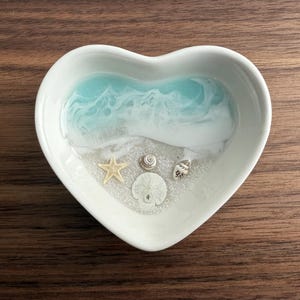 May include: A white heart-shaped dish with a blue and white resin ocean scene. The scene includes a white seashell, a starfish, and a sand dollar.