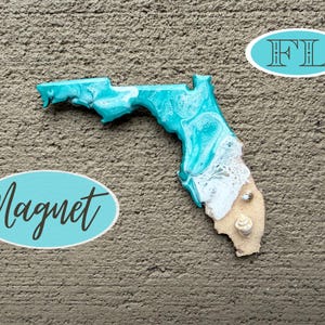 Op de afbeelding: Een magneet in de vorm van de staat Florida met een harsontwerp in turquoise en witte tinten die oceaangolven nabootsen die op een zandstrand breken, geaccentueerd met twee kleine schelpen ingebed in het getextureerde zandgedeelte.