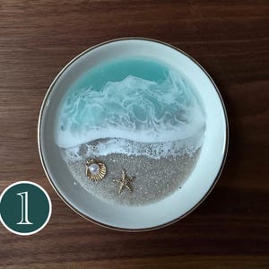 May include: A round white ceramic trinket dish featuring a resin art design of a turquoise ocean wave crashing onto a sandy beach. The beach area includes a small gold-toned seashell with a white pearl and a gold-toned starfish embellishment.