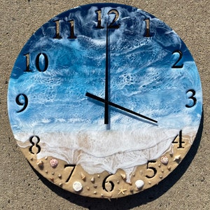 May include: A round clock with a blue and white ocean wave design. The clock face has Roman numerals and a sand-colored beach with seashells at the bottom.