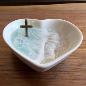 Pode incluir: Um prato de cerâmica branco em forma de coração com uma cruz dourada. O interior apresenta um design de ondas com resina azul e branca e uma seção de glitter prateado, criando uma cena de praia.