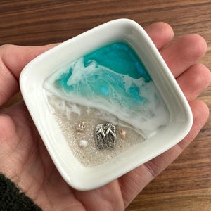 May include: A white ceramic dish with a blue and white resin design resembling a beach scene. The dish features a pair of silver flip-flops and small white seashells.