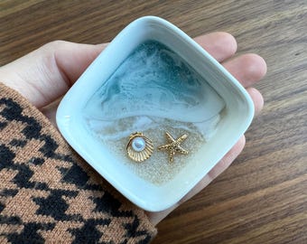 May include: A white ceramic dish with a blue and white resin design resembling a beach scene. The dish features a gold seashell with a pearl and a gold starfish.
