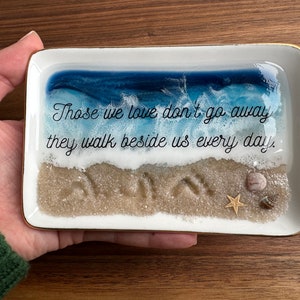 Peut inclure: Un plateau rectangulaire blanc avec une scène d'océan bleu et blanc et une plage de sable avec des empreintes de pas. Le plateau a une bordure dorée et le texte "Those we love don't go away they walk beside us every day." est imprimé sur le plateau.