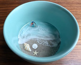 May include: A teal bowl containing a beach scene. The scene features white sand, a blue wave, a seashell, a starfish, and a small sailboat.