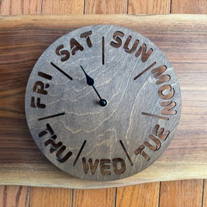 May include: A wooden clock with the days of the week cut out in a circle. The clock hands are black, and the clock is mounted on a wooden plank. The clock face is a light brown color.