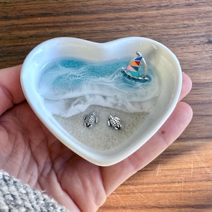 Puede incluir: Un plato de cerámica blanco en forma de corazón con una escena de playa. El plato presenta un diseño de olas azules y blancas, un velero colorido y dos pequeñas figuras de tortugas. El plato se sostiene en una mano.