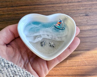 May include: A heart-shaped white ceramic dish with a beach scene. The dish features a blue and white wave design, a colourful sailboat, and two small turtle figurines. The dish is held in a hand.